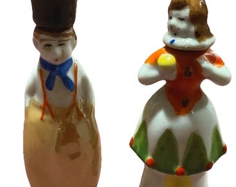 Może przedstawiać: Dwie małe figurki porcelanowe. Jedna to mężczyzna w czarnym cylindrze, niebieskiej muszce i szelkach. Druga to kobieta w białej sukience z pomarańczowymi i zielonymi akcentami. Obie są pomalowane z dbałością o szczegóły.
