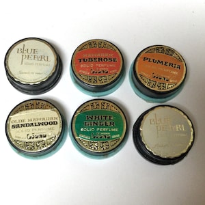 May include: Six round, black and gold compact containers of solid perfume. The containers are labeled with different floral scents: Blue Pearl, Hawaiian Tuberose, Plumeria, Olde Hawaiian Sandalwood, White Ginger, and Blue Pearl.