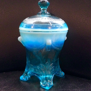 Peut inclure: Un bonbonnière en verre bleu avec un couvercle. Le plat a une base à trois pieds et des poignées. Le couvercle a un petit bouton sur le dessus. Le verre a un effet dégradé, passant d'un bleu plus clair à un bleu plus foncé.