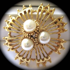 Könnte beinhalten: Eine goldfarbene Brosche mit einem filigranen, komplizierten Design, das drei weiße Perlen in der Mitte zeigt.