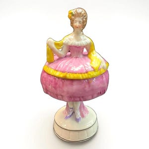 Pode incluir: Uma estatueta de porcelana de uma pessoa vestindo um vestido rosa com um xale amarelo e um acessório de cabelo amarelo. A figura está sobre uma base circular branca com um detalhe de linha preta fina. O vestido tem um corpete com babados.