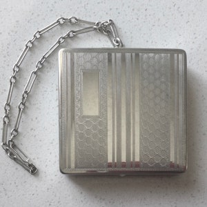 May include: A silver compact with a chain attached. The compact has a geometric pattern and a rectangular opening in the center.