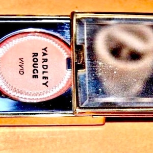 Op de afbeelding: Een gouden compact met een spiegel en twee ronde blush compacts. Een compact is gelabeld "YARDLEY ROUGE VIVID".