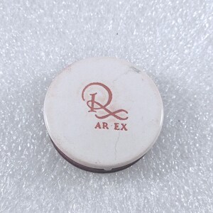 Peut inclure: Un récipient rond et blanc avec un bord marron. Le dessus présente un motif stylisé brun rougeâtre avec le texte "AR EX" en dessous. Il semble s'agir d'un produit cosmétique.