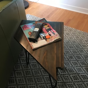 May include: A dark brown wooden triangular end table with black hairpin legs. The table has a magazine and a remote control on top of it.