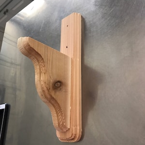 May include: A wooden shelf bracket with a decorative, curved design. The bracket is made of unfinished, light-colored wood and is ready to be stained or painted.