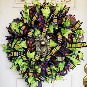 May include: A Halloween wreath with green, black, and purple ribbon, spiderweb mesh, and purple beads. A decorative metal spider is in the center of the wreath.