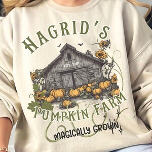 May include: Beige sweatshirt with a graphic of a barn, pumpkins, and sunflowers. Text reads "Hagrid's Pumpkin Farm" and "Magically Grown". The design is in shades of green, orange, and brown.