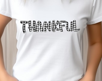Könnte beinhalten: Weißes T-Shirt mit dem Wort "THANKFUL" in großen, schwarz-weiß karierten Buchstaben auf der Brust. Das kurzärmelige Shirt scheint aus einem weichen Material zu sein. Die Person trägt eine blaue Jeans.