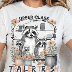 May include: White t-shirt featuring a graphic of raccoons in front of a camper, surrounded by flowers. The text reads "UPPER CLASS" above and "TRAILER TRASH" below the image. The design is in shades of gray, orange, and black.