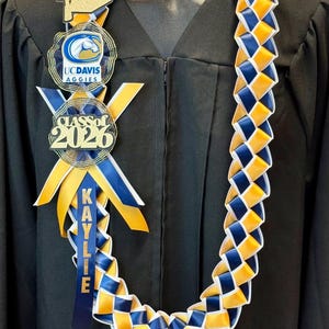 May include: A graduation lei featuring a woven pattern of blue and gold ribbons. It includes a gold glitter mortarboard, UC Davis Aggies medallions, and a "Class of 2026" emblem. A blue ribbon displays the name "Kaylie".