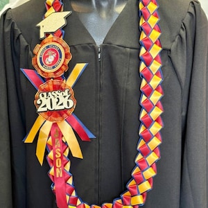May include: A graduation lei featuring red, yellow, and blue woven ribbons. It includes a gold mortarboard, a United States Marine Corps emblem, and a ribbon with "CLASS of 2026" and the name "JASON". The lei is displayed on a black graduation gown.