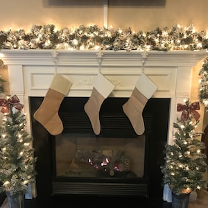 May include: A decorated fireplace with three Christmas stockings hanging from the mantel. The mantel is adorned with a garland of lights and faux snow. Two small Christmas trees flank the fireplace, each with a decorative bow.