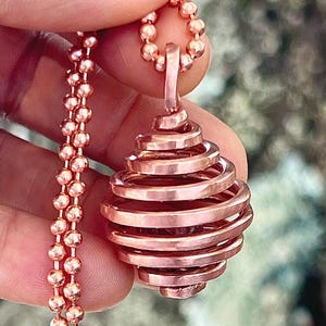 May include: A copper-colored pendant featuring a spiraled wire cage design that holds a dark stone inside. The pendant is suspended from a matching copper-colored ball chain necklace, showcasing a metallic finish and a rounded, layered structure.