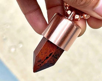 May include: A pendant necklace featuring a reddish-brown crystal with a copper-colored metal cap and chain. The crystal has a pointed, geometric shape. The chain is made of small, round beads.