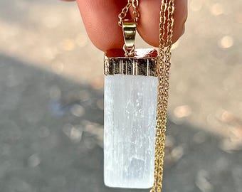 May include: A rectangular white selenite crystal pendant suspended from a thin gold-toned metal chain. The crystal has a fibrous, translucent texture and is capped with a polished gold-toned metal bail that secures it to the delicate link chain.