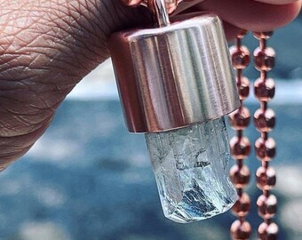 May include: A copper-colored pendant necklace featuring a cylindrical metal cap encasing a clear, crystalline gemstone. The necklace has a matching copper-colored ball chain. The gemstone has a light blue hue.