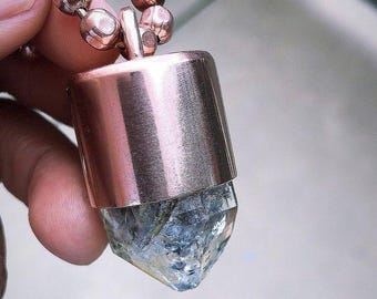 May include: A copper-colored pendant necklace with a faceted, clear crystal gemstone. The pendant has a cylindrical copper top and a crystal bottom. The necklace chain is made of small, round copper beads.