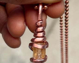 May include: A copper-colored pendant necklace featuring a cylindrical, light-orange gemstone wrapped in a spiral of copper wire. The necklace has a matching copper-colored chain. The pendant is held by a hand.