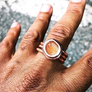 May include: A copper-colored ring with an oval, orange-colored stone. The ring has a layered band design. The stone is set in a white metal bezel. The ring is on a person's finger.