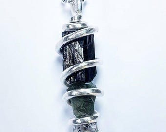 May include: A silver pendant necklace featuring a black tourmaline crystal wrapped in a spiral design. The pendant also includes a green and a gray crystal. The necklace hangs on a delicate silver chain.