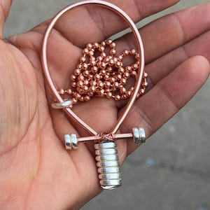May include: A copper-colored wire pendant shaped like an ankh, featuring a stack of silver-colored cylindrical beads at the base and two silver-colored beads on the crossbar. A copper-colored ball chain is looped through the top of the pendant.