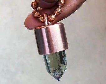 May include: A copper-colored pendant with a faceted, light blue crystal. The pendant has a cylindrical top and is attached to a copper ball chain. The crystal is pointed at the bottom.