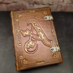 May include: A brown leather-bound journal with gold accents and a decorative gold letter "A" on the front cover. The journal has two silver clasps.