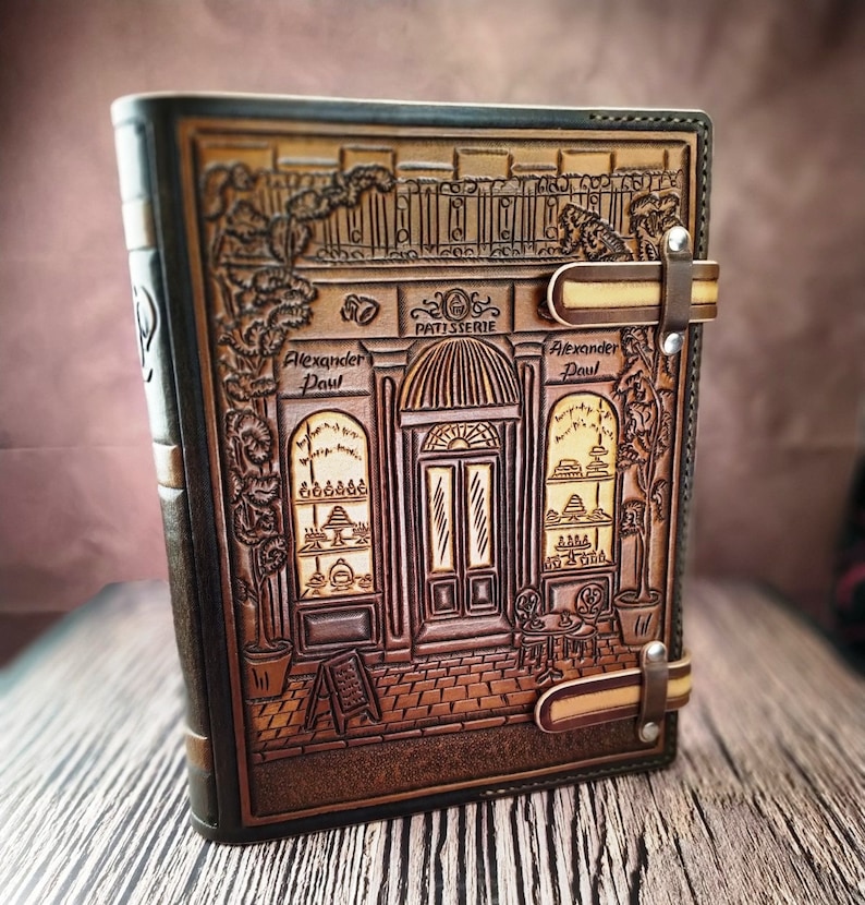 May include: A brown leather journal with a detailed embossed design of a storefront with the text "Patisserie Alexander Paul" above the door. The journal has two leather straps with metal buckles.