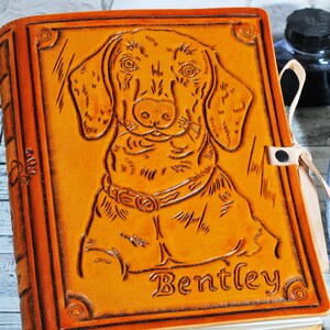 May include: A brown leather journal with a tooled image of a dachshund dog and the name "Bentley" embossed on the front. The journal has a leather strap closure and a cream-colored bookmark.