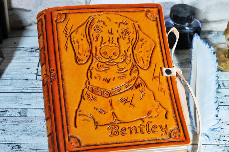 May include: A brown leather journal with a tooled image of a dachshund dog and the name "Bentley" embossed on the front. The journal has a leather strap closure and a cream-colored bookmark.