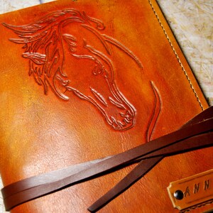 May include: A brown leather journal with a horse head design embossed on the front cover. The journal has a brown leather strap closure and a metal tag with the name "ANNA" engraved on it.