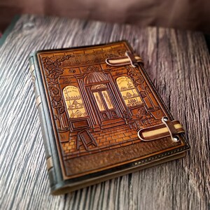 May include: A brown leather journal with a detailed embossed design of a storefront with a sign that reads "Alexander Jou" and "Natural Perfumes". The journal has two leather straps with metal buckles.