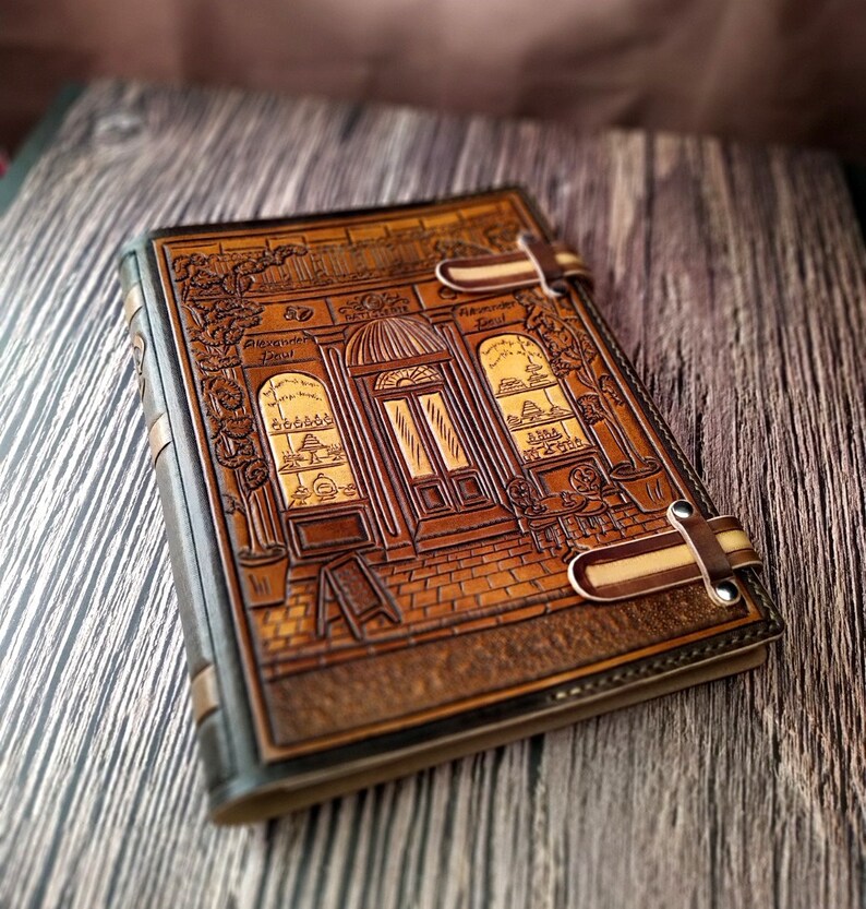 May include: A brown leather journal with a detailed embossed design of a storefront with a sign that reads "Alexander Jou" and "Natural Perfumes". The journal has two leather straps with metal buckles.