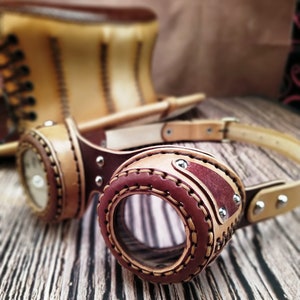 May include: Leather steampunk goggles with brown and red leather straps and a clear lens. The goggles are on a wooden surface.