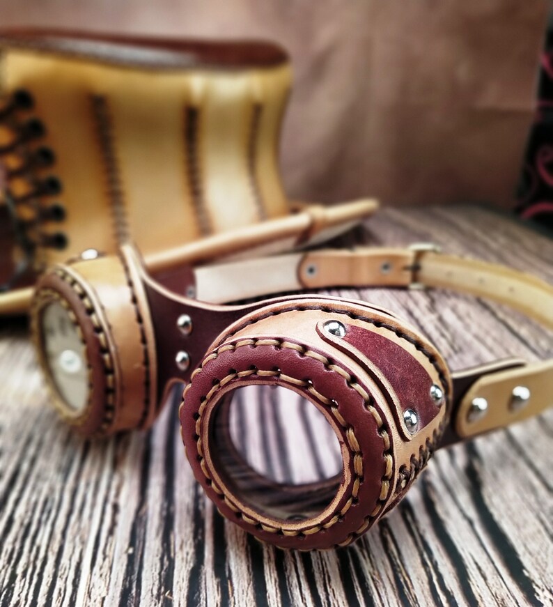 May include: Leather steampunk goggles with brown and red leather straps and a clear lens. The goggles are on a wooden surface.