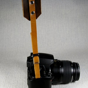 May include: A brown leather camera strap with metal hardware attached to a black DSLR camera with a lens.