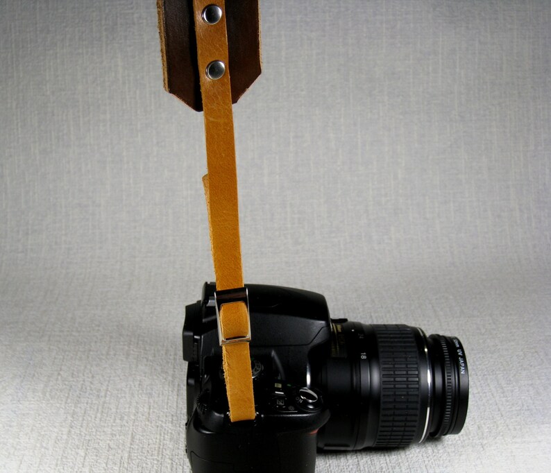 May include: A brown leather camera strap with metal hardware attached to a black DSLR camera with a lens.