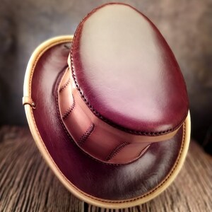 May include: A burgundy leather top hat with a tan leather brim and stitching detail. The hat is made of high-quality leather and has a classic design.