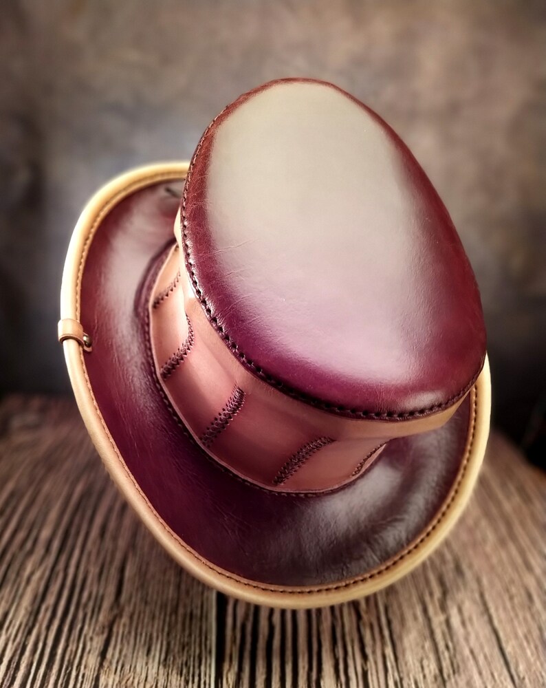 May include: A burgundy leather top hat with a tan leather brim and stitching detail. The hat is made of high-quality leather and has a classic design.