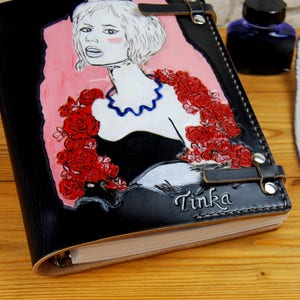 May include: Black leather journal with a hand-painted illustration of a woman with red roses and the name "Tinka" embossed on the cover.