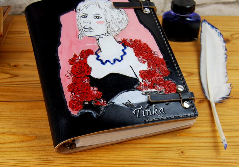 May include: Black leather journal with a hand-painted illustration of a woman with red roses and the name "Tinka" embossed on the cover.