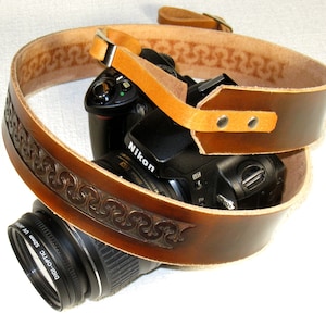 May include: A brown leather camera strap with a decorative embossed pattern. The strap is wrapped around a black camera with a lens attached. The camera has the word "Nikon" printed on it.