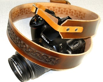 May include: A brown leather camera strap with a decorative embossed pattern. The strap is wrapped around a black camera with a lens attached. The camera has the word "Nikon" printed on it.