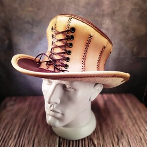 May include: A tan leather cowboy hat with a brown leather band and a brown lace-up detail on the crown.