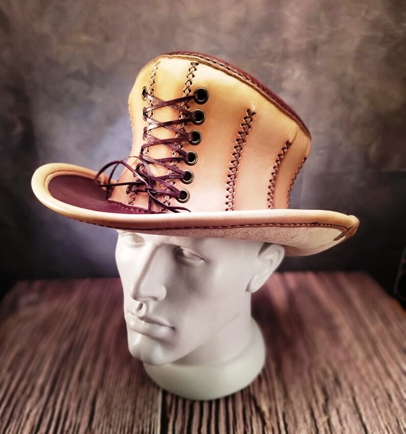 May include: A tan leather cowboy hat with a brown leather band and a brown lace-up detail on the crown.