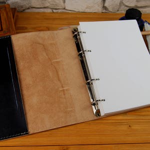 May include: Open brown leather bound journal with black leather spine and blank white pages. The journal has six silver rings for binding.