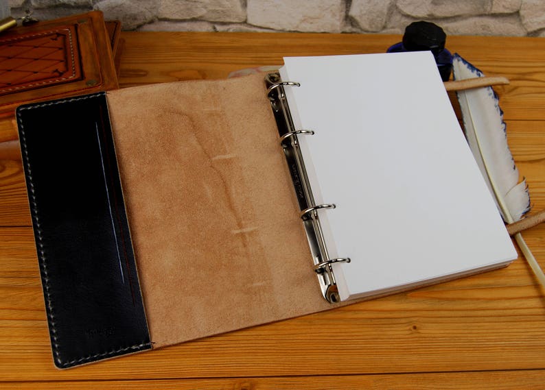 May include: Open brown leather bound journal with black leather spine and blank white pages. The journal has six silver rings for binding.
