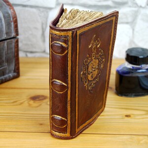 May include: A brown leather-bound journal with gold accents and a decorative design on the front cover. The journal is closed and standing upright on a wooden surface.