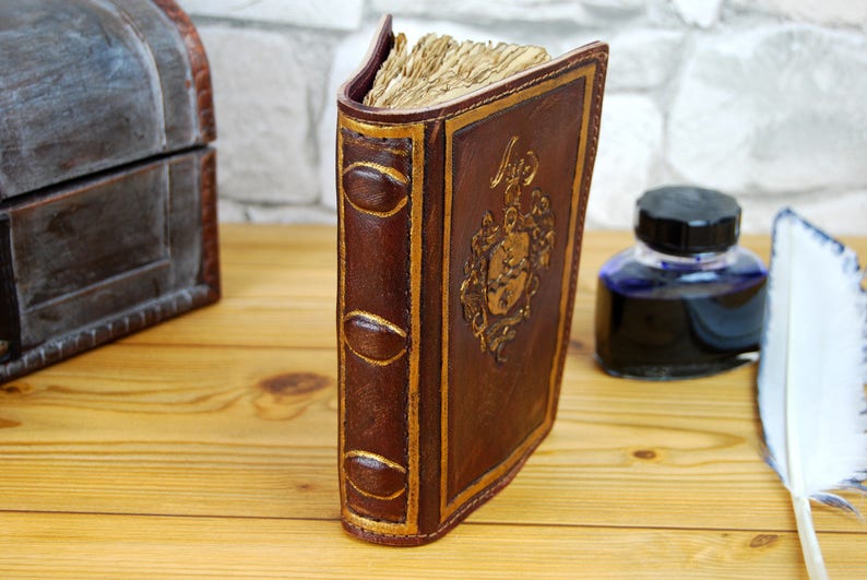 May include: A brown leather-bound journal with gold accents and a decorative design on the front cover. The journal is closed and standing upright on a wooden surface.